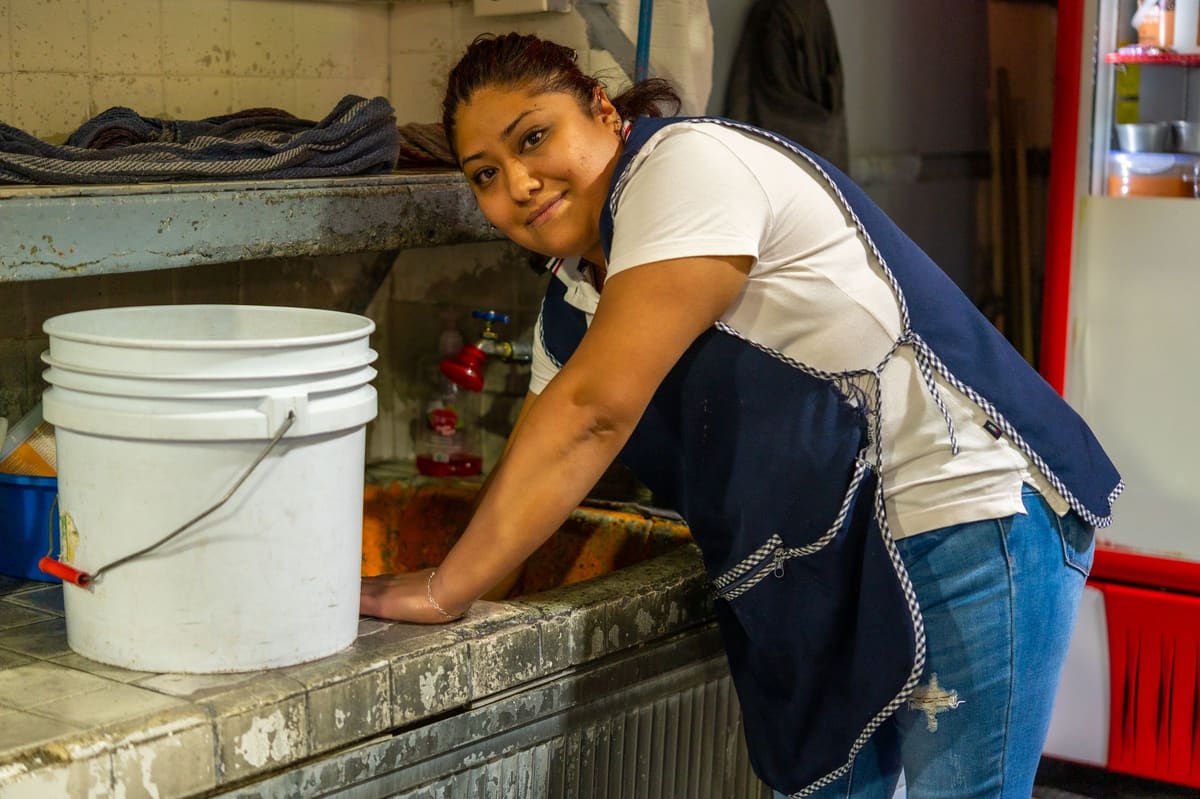 Mujer con delantal sonriendo mientras lava una cubeta en una pila